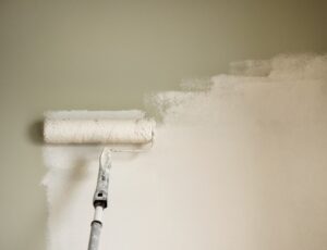 Roller applying a coat of fresh paint to an interior wall, covering an olive-green surface with a lighter neutral color, with the painted and unpainted sections clearly visible