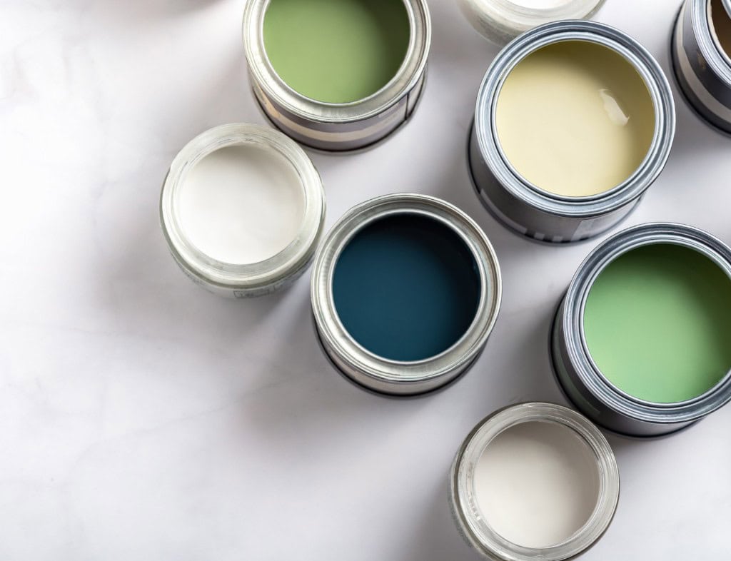 Open paint cans in green, blue, yellow, and white arranged on a white surface, showing interior paint color and sheen options like flat and high-gloss.